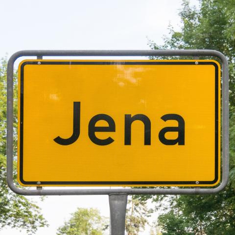 Ein gelbes Ortseingangsschild mit dem Schriftzug Jena vor einem Naturhintergrund