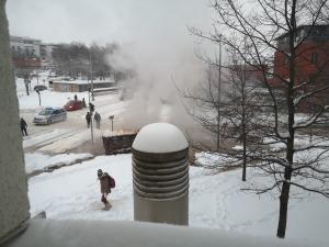 Blick auf eine verschneite Straße mit einer Rauch- bzw. Dampfwolke, mehrere Fahrzeuge, Fußgänger und eine Polizeiauto