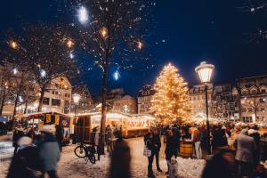 Am Abend, es schneit: Im Hintergund Häuser des Jenaer Marktes, Weihnachtsmarktstände der beuchtete Weihnachtsbaum. Im Vordergund Menschen.