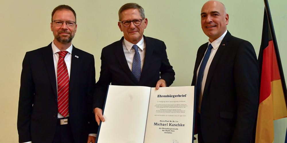 Drei Männer stehen nebeneinander, einer hält ein großes Buch in de Hand.