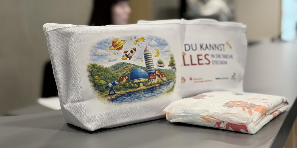 Zwei weiße Stofftaschen mit buntem Aufdruck stehen auf einem Tisch, daneben liegen Windeln; im Hintergrund unscharf Personen in einem Innenraum.