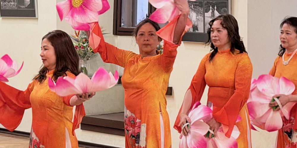 Mehrere Frauen in orangefarbenen traditionellen Kleidern führen einen Tanz mit großen rosa Lotusblüten in einem hellen Ausstellungsraum auf.
