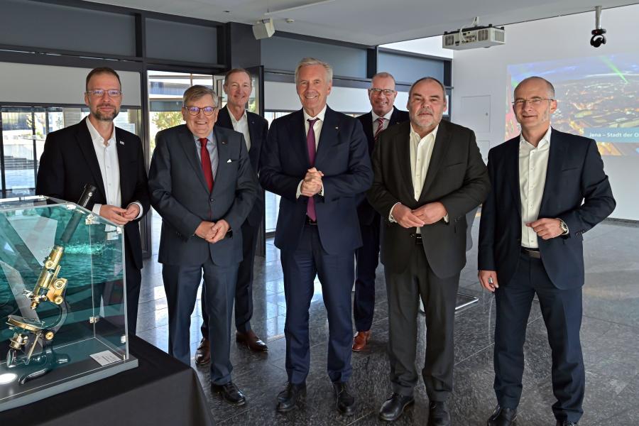 Sieben Männer stehen nebeneinander in einem Besprechungsraum, im Vordergrund ist ein historisches Mikroskop zu sehen