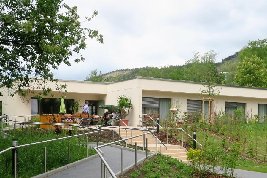 Blick auf eine flaches helles Gebäude, im Vordergrund eine Treppe und eine Rampe die durch einen Garten zum Haus führt.