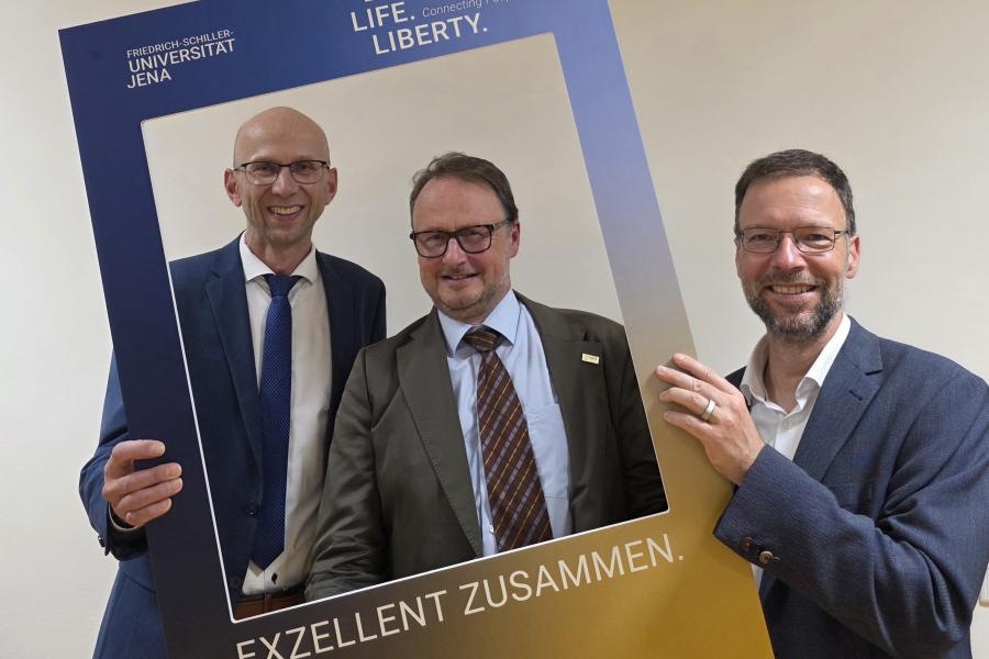 Drei Männer halten einen Aufsteller in Form eines Polaroid-Bildes und schauen hindurch. Auf dem Schild steht "Exzellent zusammen"