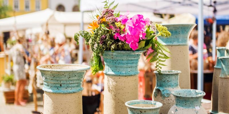 Blau-beige Keramiken, in einer Vase davon steckt ein Blumenstrauß, im Hintergrund Menschen & Stände auf dem Jenaer Töpfermarkt