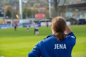 Trainerin mit Trainigsjacke mit der Aufschrift Jena, im Hintergund ein Fussballfeld mit Spielerinnen