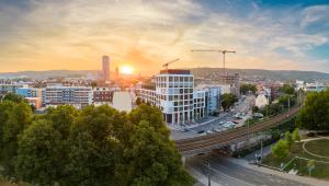 Blick auf eine Stadt mit mehreren großen Gebäuden und Türmen, einer Bahnstrecke, viel Grün und einem Sonnenuntergang am Horizont