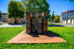 Denkmal mit mehren Türmen von Aktenbehältern in unterschiedlichen Farben und Aufschriften, steht auf einer grünen Wiese. Im Hintergrund sind Bäume und andere Gebäude zu sehen.