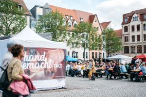 Besucher:innen schlendern über den Jenaer Markt zu den handgemacht-Tagen an einem Pavillon mit dem Titel der Veranstaltung entlang, im Hintergrund sitzen weitere Gäste.