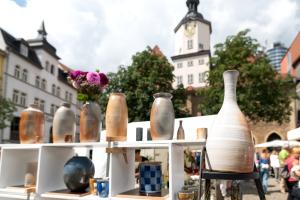 Blick auf den Jenaer Töpfermarkt mit Historischem Rathaus und Jentower, im Vordergrund Vasen in einem Regal, im Hintergrund tummeln sich Besucher:innen an den Ständen