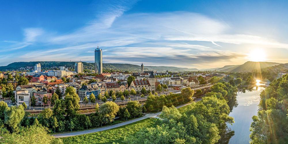 Blick aus der Vogelperspektive auf eine Stadt mit einem Fluss, einer Eisenbahntrasse, Gebäuden, viel Grün und Bergen im Hintergrund