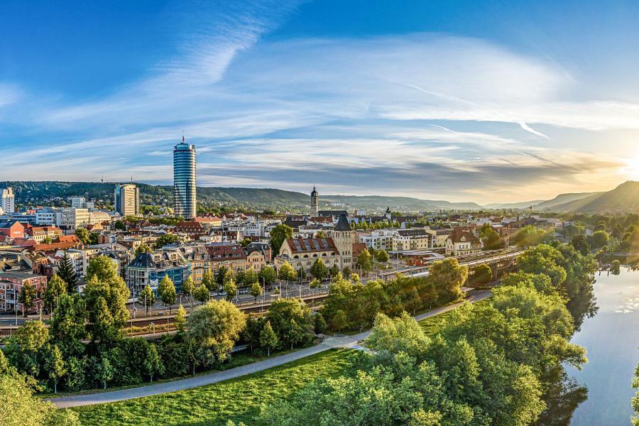 Blick aus der Vogelperspektive auf eine Stadt mit einem Fluss, einer Eisenbahntrasse, Gebäuden, viel Grün und Bergen im Hintergrund