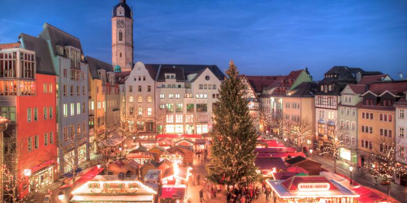 Blick aus der Vogelperspektive auf einen weihnachtlich beleuchteten Marktplatz mit einem großen Baum in der Mitte und vielen Ständen und Menschen