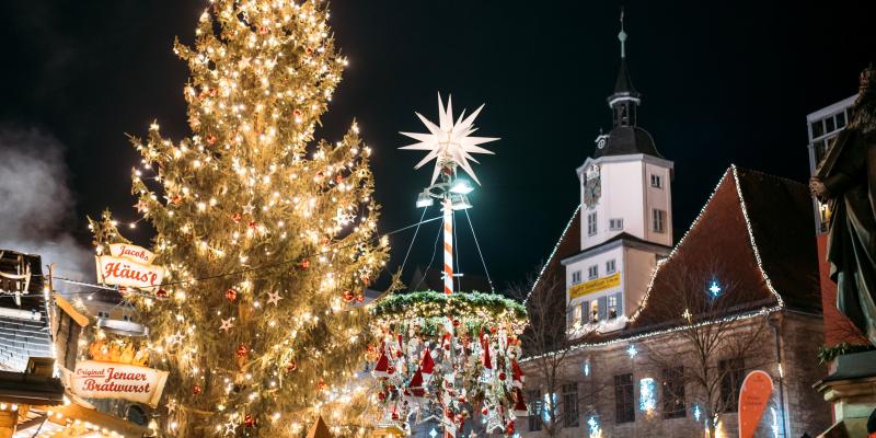 Blick auf den Jenaer Weihnachtsmarkt zu abendlicher Stunde, mit Weihnachtsbaum im Vordergrund, im Hintergrund Stände